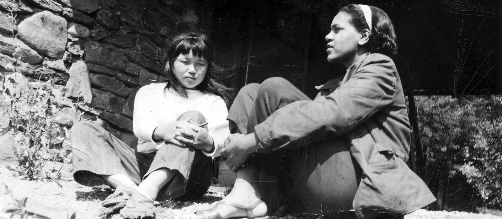 Black and white photograph of a young woman with black hair and asian features wearing a white top and medium shaded pants crouched next to a young woman with black hair pulled back with a light colored headband with darker skin, jacket, and pants, hugging her knees to her body with stacked rocks and darkness in the background